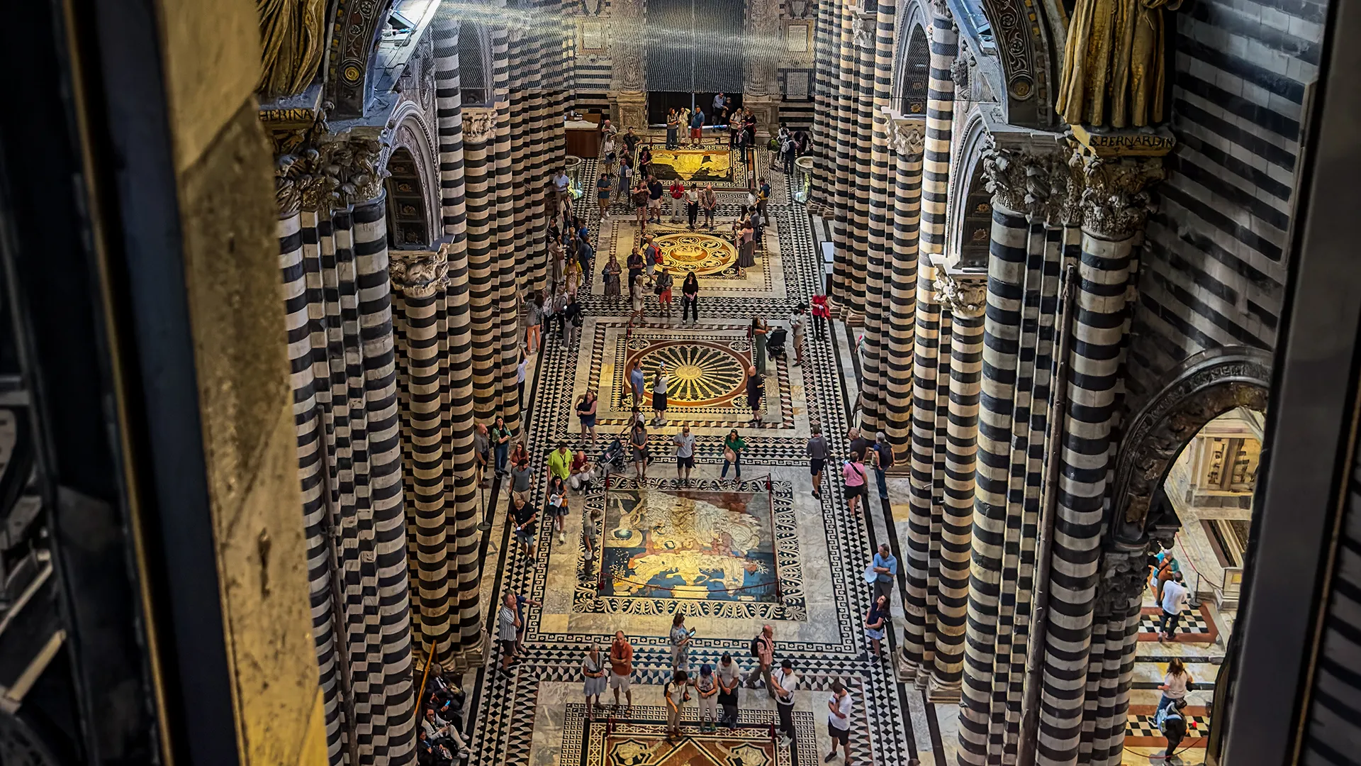 Inlaid marble floor of the Duomo di Siena seen from above between the cathedral’s striped columns