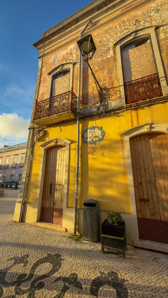 Weathered yellow building with wooden doors, iron balconies, and decorative calçada on Rua 5 de Outubro in Loulé, Portugal.