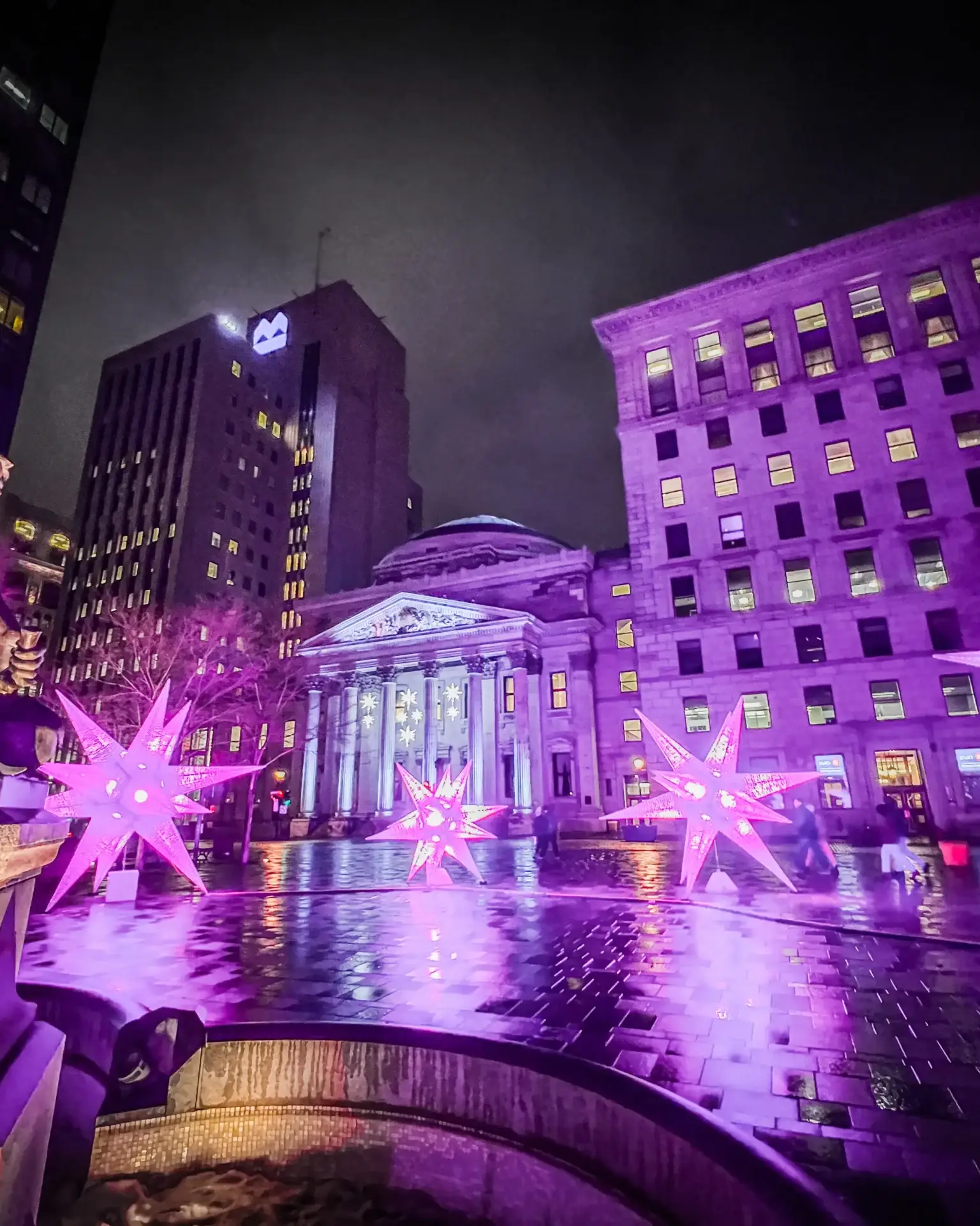 Pink and purple illuminated stars reflected on wet pavement at Place d’Armes in Old Montréal, with a sculpted fountain on the left and historic buildings behind.