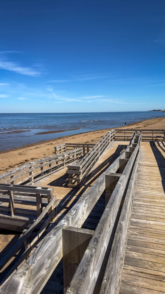 Wooden ramp with switchbacks and railings descending toward Aboiteau Beach, with sand and water beyond.
