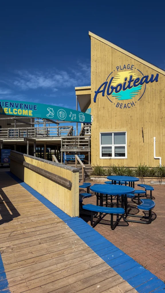 Aboiteau Beach sign on the main pavilion beside a bilingual welcome banner, wooden walkways and empty blue picnic tables.