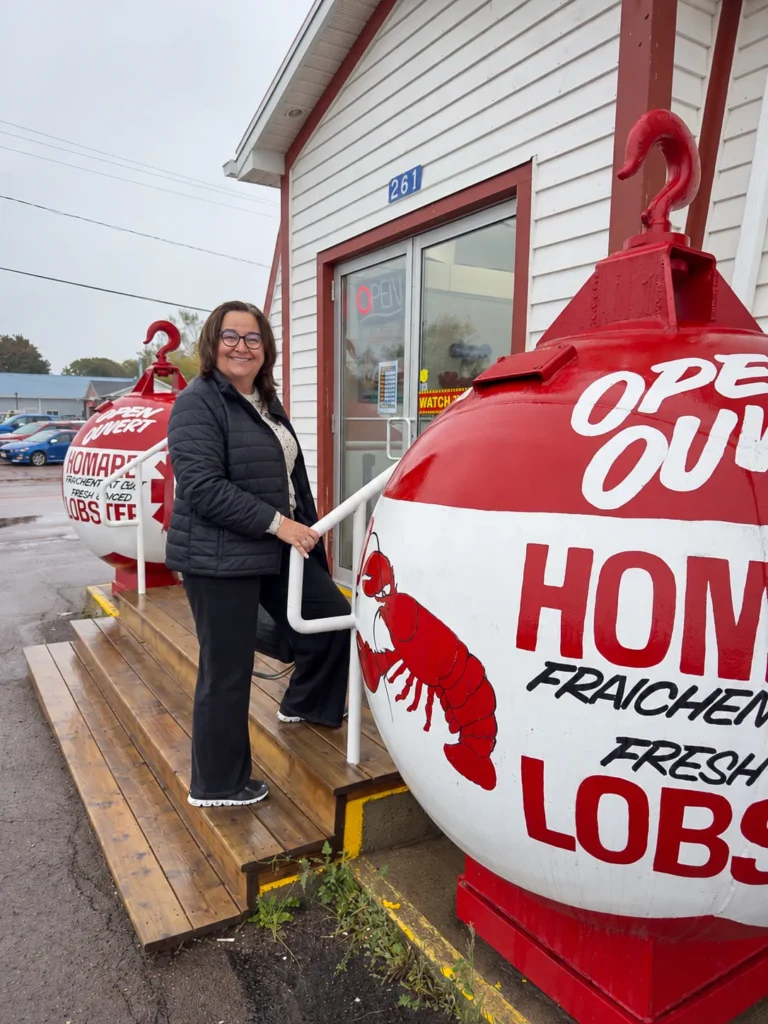 Danielle standing on wooden steps outside the lobster shop, between two large red-and-white lobster buoys.