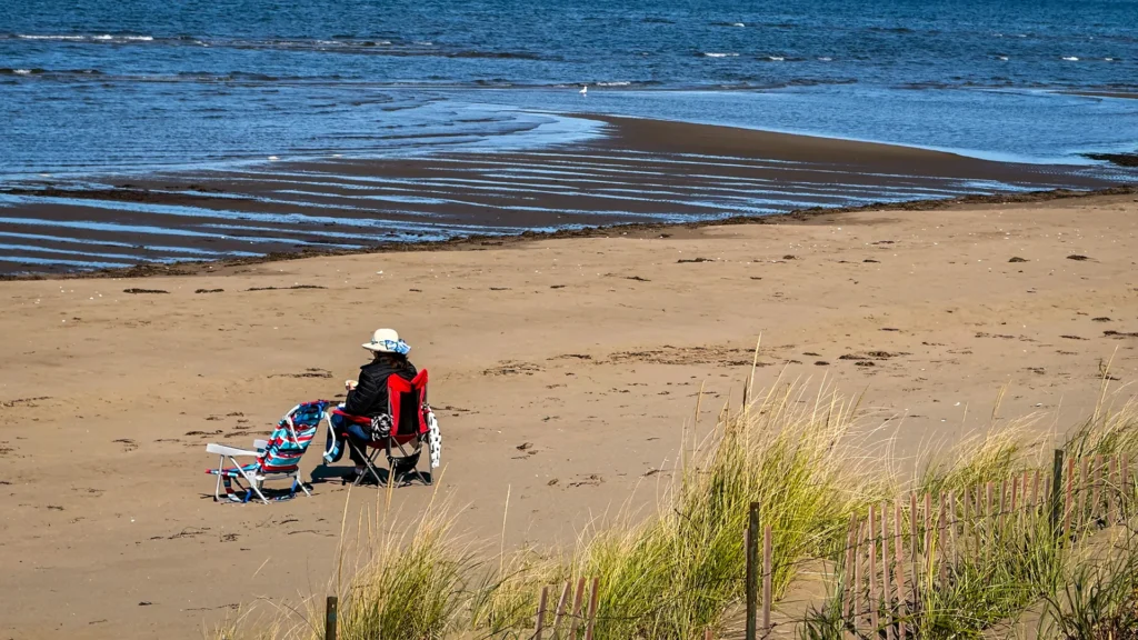 Person sitting in a beach chair on a broad sandy shore, with dune grasses in the foreground and shallow water beyond.