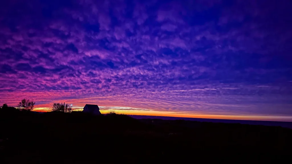 Purple and magenta clouds above an orange horizon, with a house and trees in silhouette.