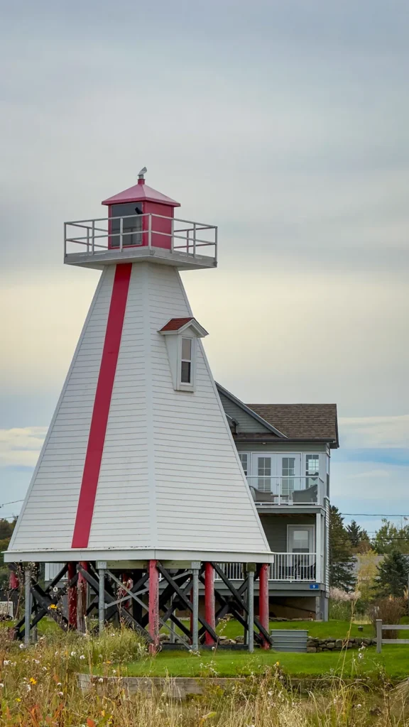 Red-and-white lighthouse on wooden supports, with a house behind it and grasses in the foreground.