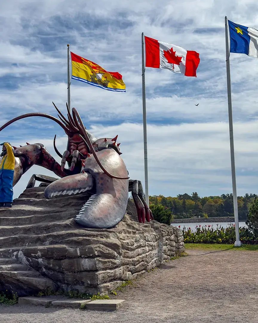 Giant lobster sculpture beside the water, with a visitor posing near its claw and three flags flying overhead.